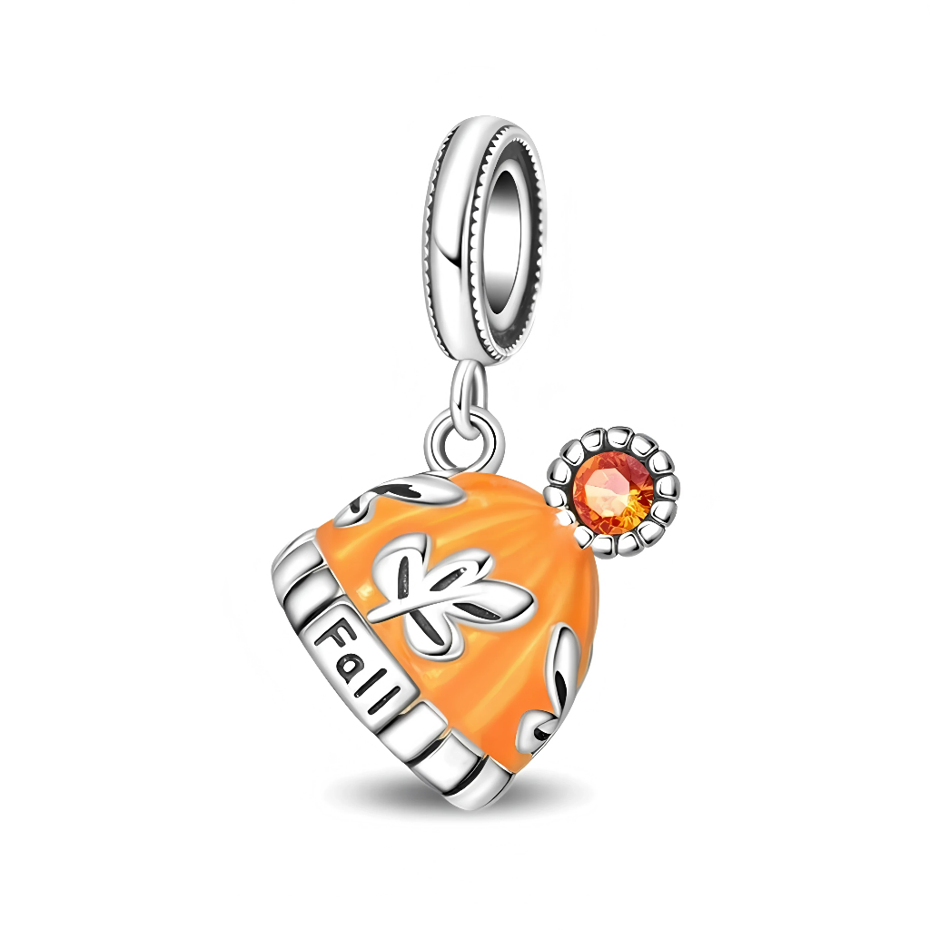 La bélière en argent 925 présente un bord perlé discret et laisse pendre un bonnet émaillé orange, ajouré de motifs de feuilles argentées sur les côtés. Sur le bord du bonnet est gravé le mot « Fall », tandis qu’un petit médaillon rond serti d’un cristal orange vif rappelle la lumière dorée des journées d’automne.