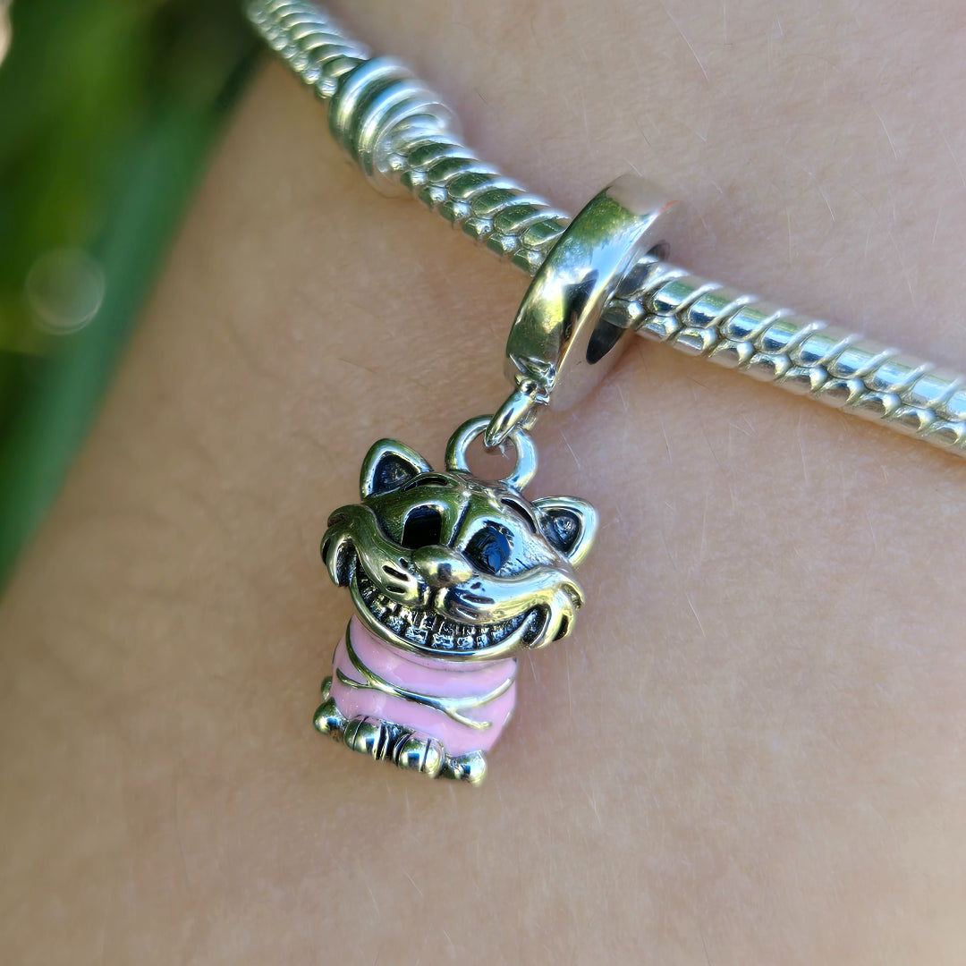 Ajoutez une touche de folie et de mystère à votre collection avec ce charm pendentif du célèbre Chat du Cheshire, issu de l’univers d’Alice au Pays des Merveilles. Assis dans sa posture emblématique, ce personnage malicieux est ici représenté en finition argentée brillante, avec un corps rose rayé fidèle à son design original. Son sourire énigmatique et son regard espiègle font de ce bijou un clin d'œil parfait à l'imaginaire décalé de Lewis Carroll.