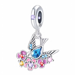 Ce charm célèbre la légèreté et la couleur. Le petit oiseau stylisé arbore des ailes ponctuées de micro‑cristaux bleus et un corps sublimé par une pierre centrale bleu azur taille poire. Il survole un parterre de fleurs roses, violettes et jaunes rehaussées de cabochons scintillants. La bélière est sertie d’une gemme lavande qui renforce l’harmonie pastel. Idéal pour un thème nature, bohème ou renouveau.